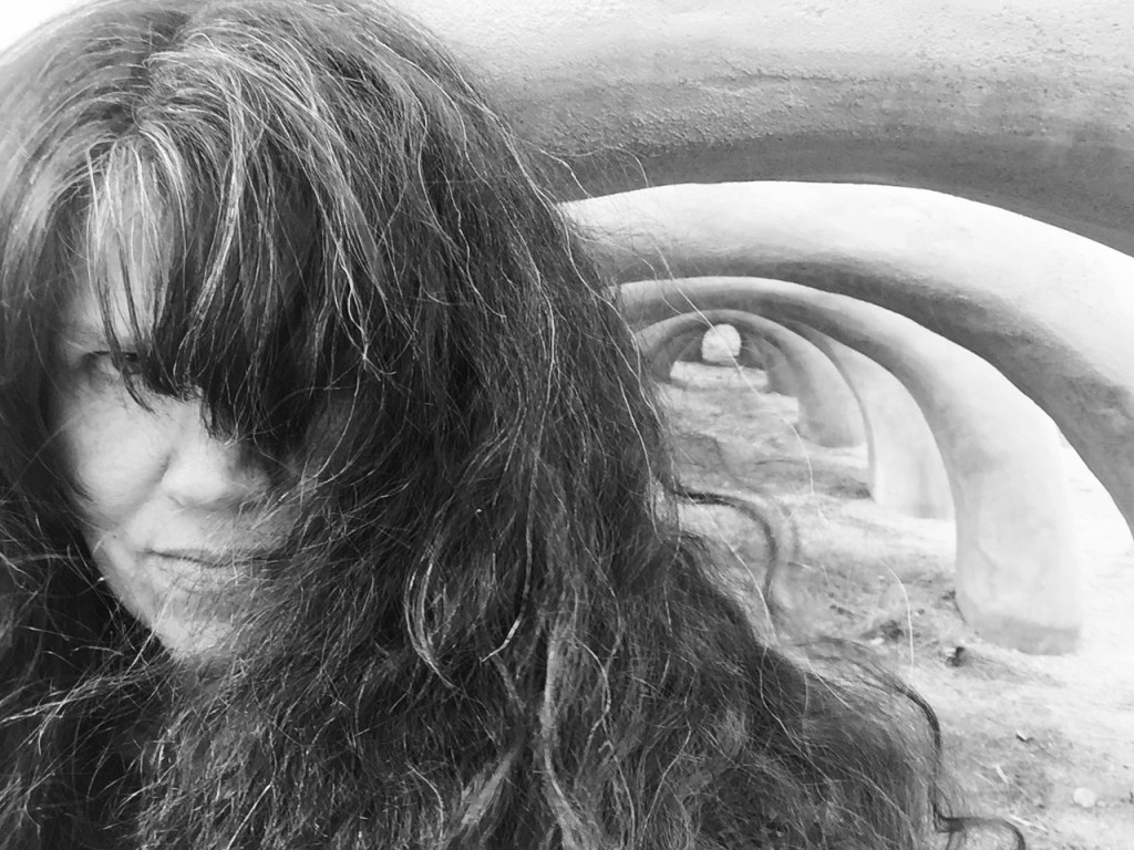 Black & White selfie of the author with her hair blowing in front of most of her face, with cool arches in the background.